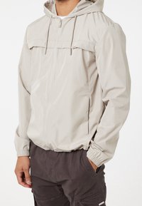 Ljust beige zip-jacka med huva, elastiska ärmslutar och en framficka. Har ett mjukt, vattenavvisande material och en minimalistisk design.