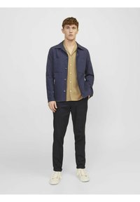Giacca strutturata blu navy con chiusura a bottoni, due tasche a toppa, indossata sopra una camicia col beige. Pantaloni neri e sneakers color crema completano l'outfit.
