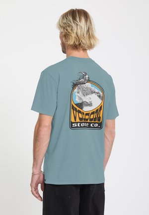Homme aux cheveux blonds portant un T-shirt bleu avec un graphisme de surfeur chevauchant une vague et le texte « Volcom Stone Co. » au dos, vu de dos.