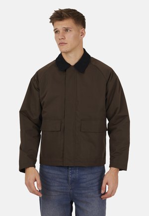 Młody mężczyzna w ciemnobrązowej kurtce z czarnym kołnierzem i przednimi kieszeniami na klapę, stojący na jednolitym jasnym tle.