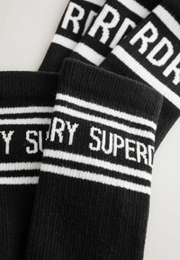 Juodų mezginių kojinių su baltomis juostelėmis ir užrašu "SUPERDRY". Audinys turi gilią tekstūrą.
