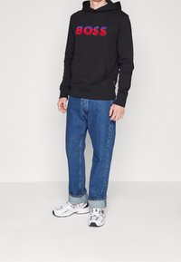 Felpa nera con cappuccio con logo "BOSS" rosso e blu, abbinata a jeans azzurri e sneaker bianche con dettagli neri.