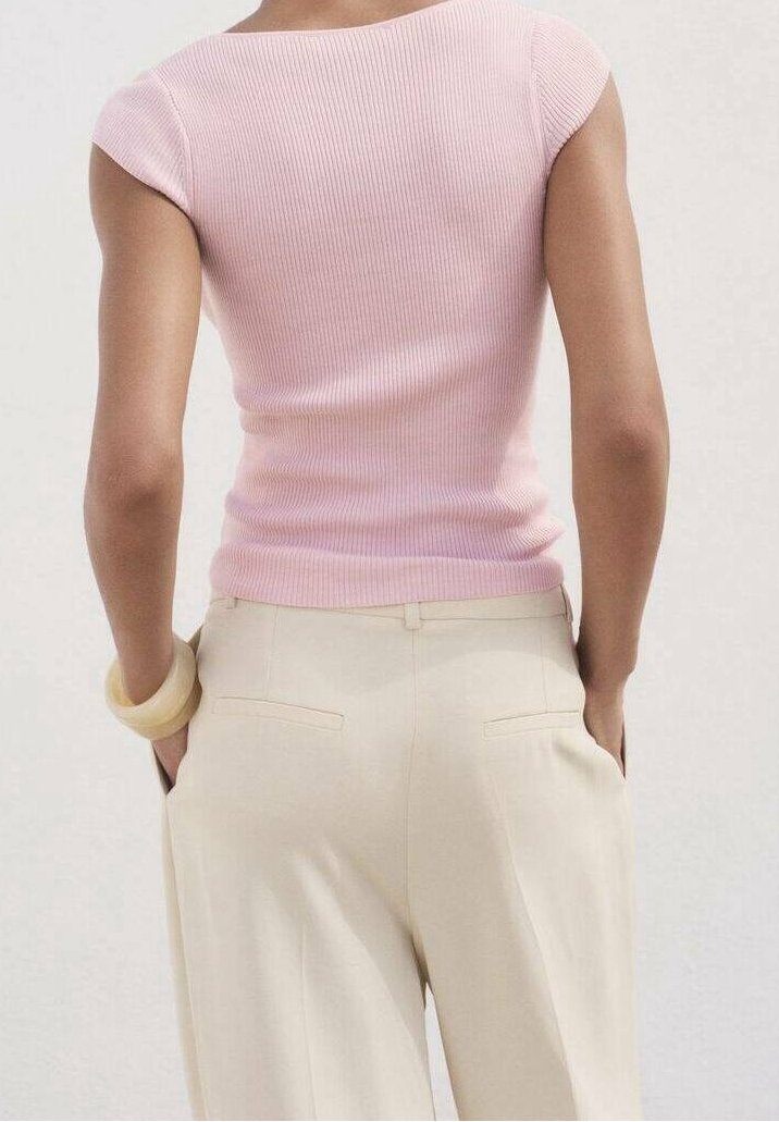 Personne portant un haut rose ajusté à manches courtes et un pantalon beige taille haute, debout les mains dans les poches devant un fond uni.