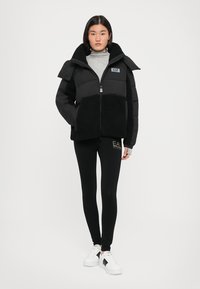 Zwarte pufferjack met een contrasterende fleece onderkant, hoge kraag, ritssluiting en logo. Gepaard met zwarte legging en witte sneakers.
