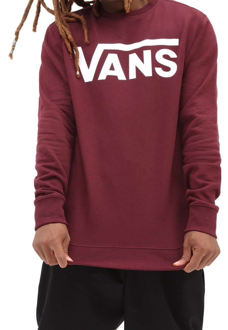 Personne portant un sweat Vans bordeaux avec un logo blanc, tenant l'ourlet, montrant les manches longues et un pantalon noir.