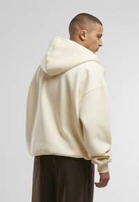 Cremefarbener Kapuzenpullover mit lockerem Schnitt, großer Kapuze, gerippten Bündchen und weichem Material, kombiniert mit braunen Hosen.