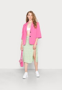 Blazer oversized em rosa brilhante sobre um top de crop branco, combinado com uma saia floral verde-claro com uma fenda lateral. Ela segura uma bolsa rosa e usa tênis brancos.