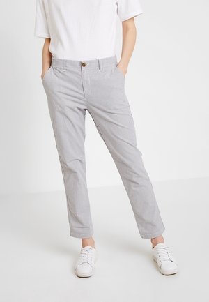 Personne portant des baskets blanches et un pantalon gris clair à fines rayures, les mains dans les poches, debout sur un fond blanc.