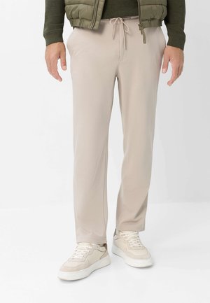 Uomo che indossa pantaloni beige con coulisse, sneakers beige, maglione verde e giacca trapuntata verde oliva, in piedi su pavimento bianco.