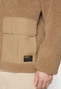 Chaqueta de fleece marrón con un panel de tela color crema y un bolsillo con parche. Incluye una etiqueta negra con texto amarillo, costuras visibles y tela texturizada.
