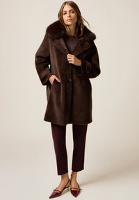 Kobieta ubrana w ciemnobrązowy płaszcz z sztucznego futra z szerokim kołnierzem, bordowe spodnie i bordowe szpilki, stojąca na jednolitym tle.