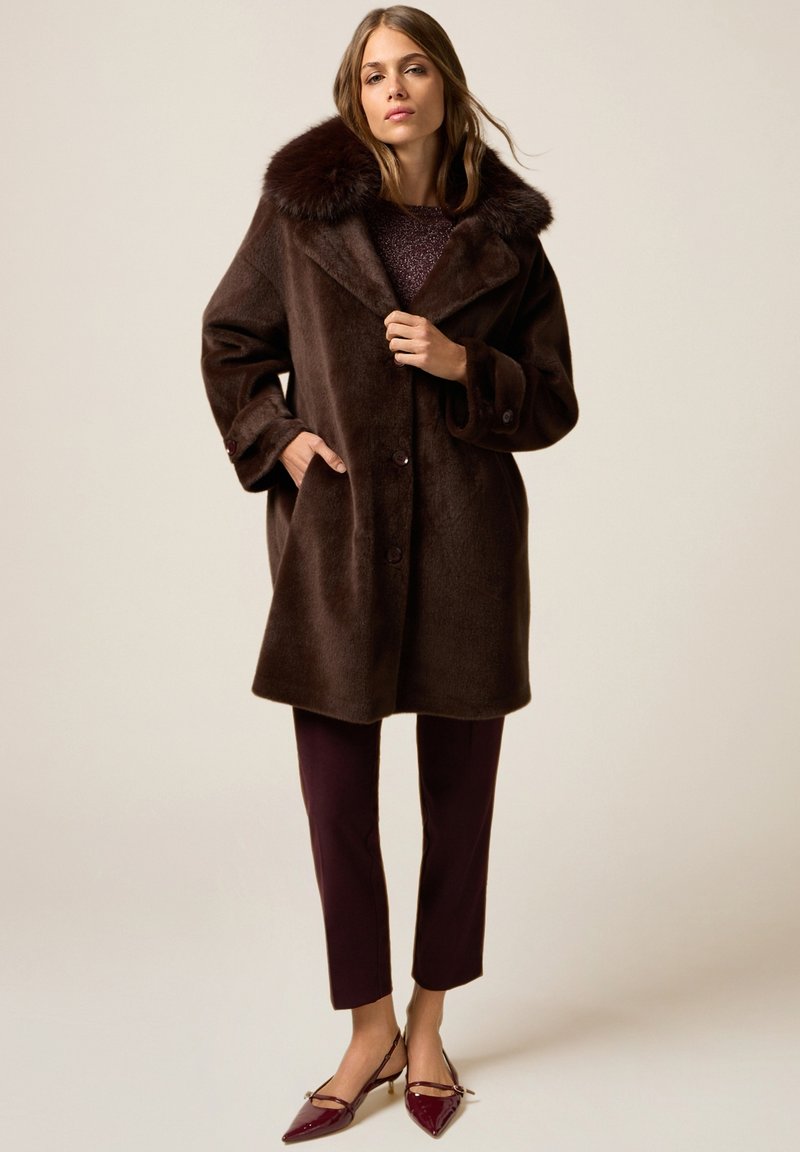 Kobieta ubrana w ciemnobrązowy płaszcz z sztucznego futra z szerokim kołnierzem, bordowe spodnie i bordowe szpilki, stojąca na jednolitym tle.