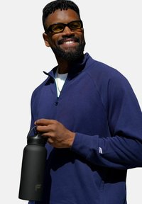 Schwarze isolierte Wasserflasche, die in der Hand gehalten wird, mit mattem Finish. Das Modell trägt einen navyfarbenen Reißverschluss-Pullover mit hohem Kragen und gerippten Bündchen.