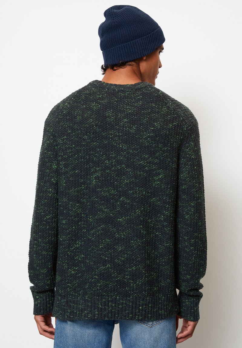 Pull en tricot de couleur vert foncé avec des taches vertes plus claires, présentant un col et des poignets côtelés. Porté avec un bonnet tricoté bleu.