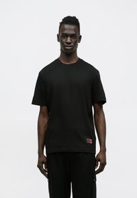 Μαύρο βαμβακερό T-shirt με στρογγυλή λαιμόκοψη και κοντά μανίκια. Διαθέτει κόκκινη ετικέτα στο κάτω αριστερό μέρος με το κείμενο "HUGO" σε έντονη γραφή.
