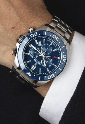 Montre chronographe en acier inoxydable avec un cadran bleu, des index horaires lumineux et des accents argentés. Elle est équipée d'un tachymètre et d'un affichage de la date.