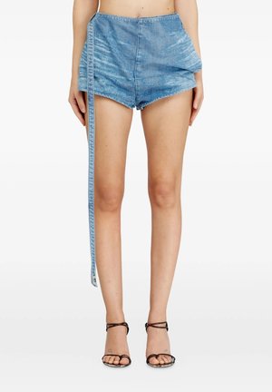 Modella con shorts di jeans azzurri a vita alta con una fascia estesa sul lato sinistro, abbinati a sandali neri con tacco alto e cinturini.