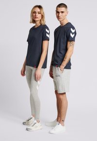 En marinblå t-shirt med vita chevron-accenter, kombinerad med grå leggings med liknande accenter. Grå shorts med logotyp, bärs med vita sneakers.