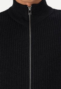 Maglione nero lavorato a maglia con un colletto alto e una zip frontale. La texture è a costine, offrendo un aspetto accogliente. Dettaglio della zip metallica visibile.