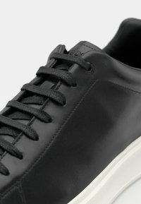 Černá kožená teniska s texturovanými černými tkaničkami, hladkým povrchem a bílou gumovou podrážkou. Minimalistický design bez výrazných vzorů.