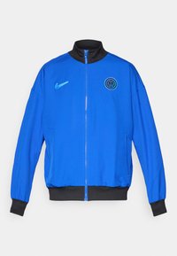 Blå Nike jakke med sort krave, lynlås foran og ribbede sorte manchetter. Har Nike-logoen og et cirkulært holdemblem på brystet.