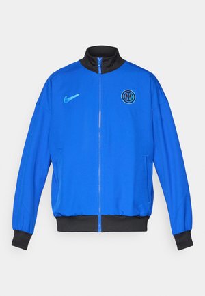 Modrá Nike bunda s čiernym golierom, zipsom vpredu a ribbovanými čiernymi manžetami. Obsahuje logo Nike a okrúhly tímový erb na hrudi.