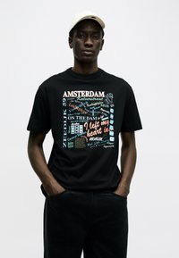 Junger Mann trägt eine cremefarbene Baseballkappe und ein schwarzes T-Shirt mit buntem, Amsterdam-inspiriertem Schriftzug und Grafik, Hände in den Hosentaschen, neutraler Hintergrund.