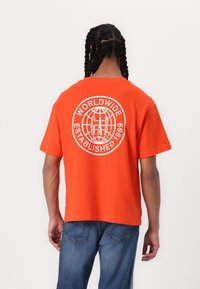 Oranje katoenen t-shirt met een ronde hals, korte mouwen en een grote witte grafische afdruk op de achterkant met een wereldbol en merktekst.