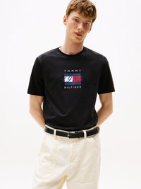 T-shirt en coton noir avec un design graphique portant "TOMMY HILFIGER" et l'année "1985" en rouge, blanc et bleu. Porté avec un pantalon crème et une ceinture noire.