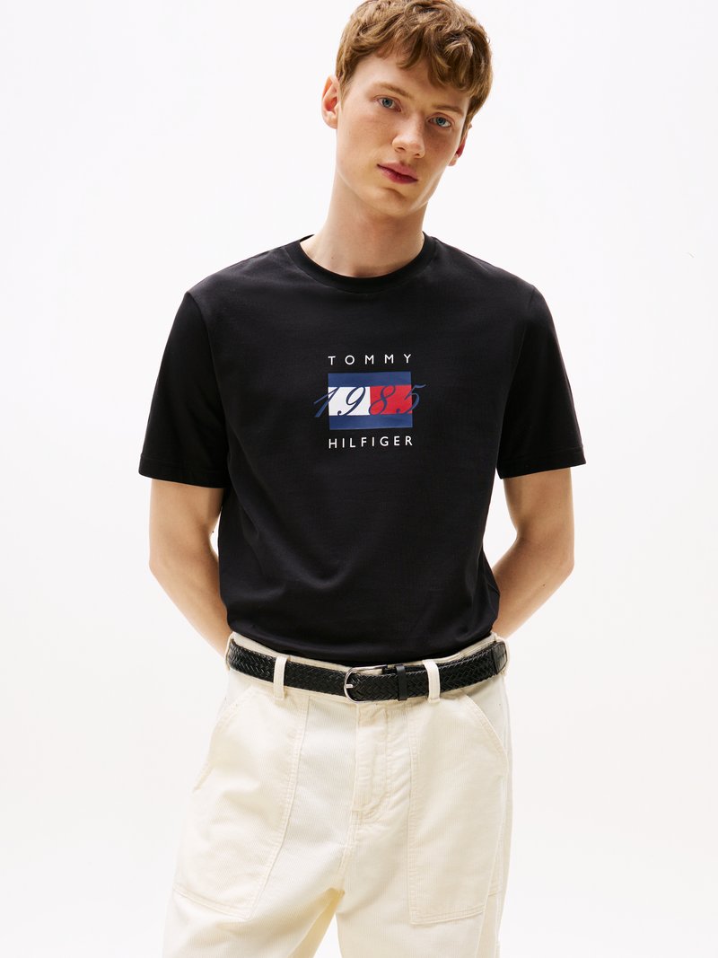 T-shirt en coton noir avec un design graphique portant "TOMMY HILFIGER" et l'année "1985" en rouge, blanc et bleu. Porté avec un pantalon crème et une ceinture noire.
