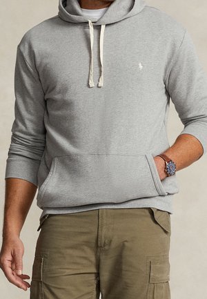 Mann trägt hellgrauen Hoodie mit weißen Kordeln, hellbraune Cargohose und eine Armbanduhr mit braunem Lederarmband am linken Handgelenk.