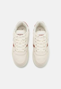 Sneakers Lacoste in tela crema con righe di accento marrone. Presentano lacci tondi, collo imbottito e suola in gomma. Design minimalista.