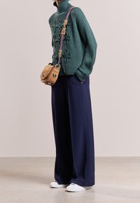 Groene gebreide trui met hoge kraag en decoratieve strikdetails, gecombineerd met brede marineblauwe broeken en witte sneakers. Kleine tan schoudertas.