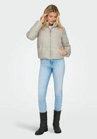 Svijetlosiva puffer jakna sa zipom sprijeda, visokim kragnom i bočnim džepovima, nosi se preko bež majice i uparena je sa svijetloplavim trapericama i crnim čizmama.