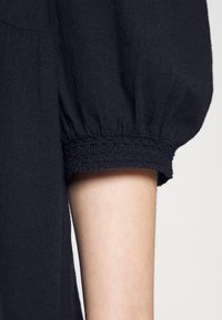 Vêtement noir à manches bouffantes avec élastique au poignet, présentant une finition texturée. Le tissu semble léger et légèrement froissé.