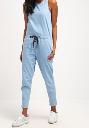 Combinaison bleu clair sans manches avec une coupe décontractée et un cordon de serrage noir réglable à la taille, associée à des baskets blanches.