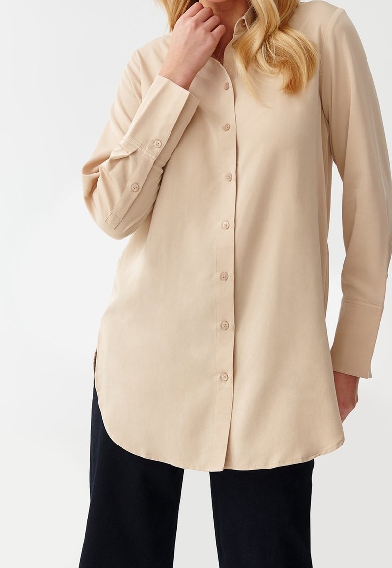 Femme portant un chemisier beige à manches longues boutonné avec un pantalon noir, main touchant le menton, sur un fond blanc uni.