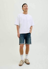 Jovem de pé com as mãos nos bolsos, vestindo uma t-shirt oversized branca, calções de ganga dobrados, meias brancas com logótipos e ténis bege.