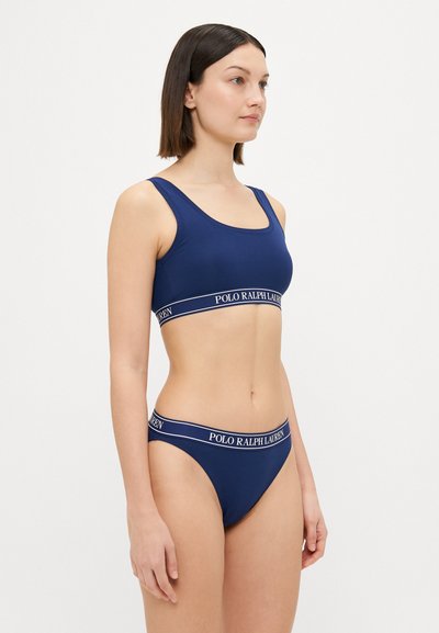 Tamsiai mėlynas sportinis liemenukas ir maudymosi kostiumėlis, turintys platų elastingą juostą su "Polo Ralph Lauren" ženklu, griovelių tekstūrą ir prigludusį siluetą.