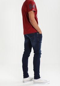 Röd kortärmad skjorta med ett blått nummer på ärmen, ihop med mörkblå slim-fit jeans och vita sneakers, med en klassisk design.