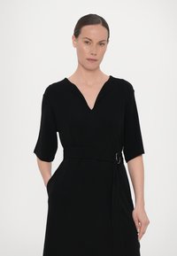 Crna haljina s V izrezom, kratkim rukavima i strukom s remenom koji ima srebrnu kopču. Glatka tkanina s elegantnim, sofisticiranim dizajnom.