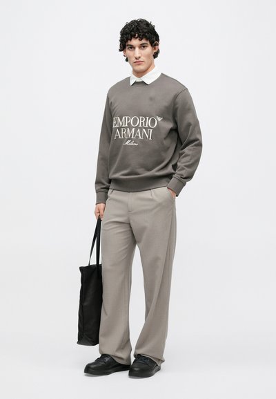 Šedá mikina s bielym logom, skombinovaná so svetlosivými nohavicami s pruhmi. Čierna taška a topánky dopĺňajú outfit. Model stojí vzpriamene.