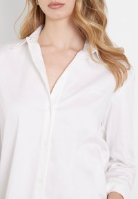 Camisa blanca de botones, hecha de algodón, con una textura suave. Presenta un pequeño logo bordado en el pecho y un diseño clásico de cuello.