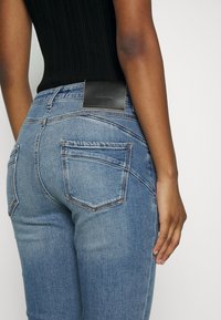 Žena v světle modrých džínách a černém topu bez rukávů, s viditelnou značkovou etiketou na pase, stojící na bílém pozadí.