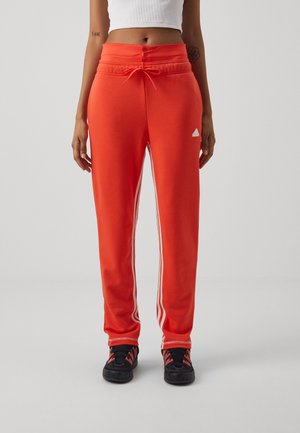 Pantaloni sportivi rossi con strisce laterali bianche, vita elastica con cordino e un piccolo logo bianco sulla parte sinistra dell'anca. Il tessuto sembra morbido.