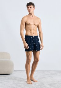 Mænds badebukser i marineblåt stof, med et mønster af små ikoner, elastisk talje og frontknaplukning.
