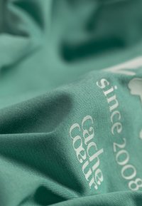 Turquoise stof met ribbeltextuur, met witte bedrukte tekst: "Cache Coeur sinds 2008." Zachte, gedrapeerde uitstraling.