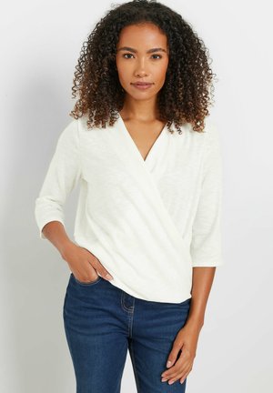 Kobieta o kręconych włosach ubrana w biały top z dekoltem w serek i rękawami do łokcia oraz ciemnoniebieskie dżinsy, stojąca z jedną ręką w kieszeni.