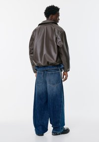 Hnedá umelá kožená bunda s voľným strihom a zníženými plecami, skombinovaná s oversized modrými džínsami s na Gathered pásom a reťazovým detailom.