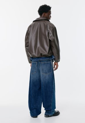 Hnědá koženka s volným střihn a spadlými rameny, spárovaná s oversized modrými džíny s nařaseným pasem a řetězovým detailem.